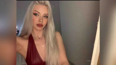 Blonde woman in a deep red silk dress posing with elegant makeup in a soft-lit indoor setting.