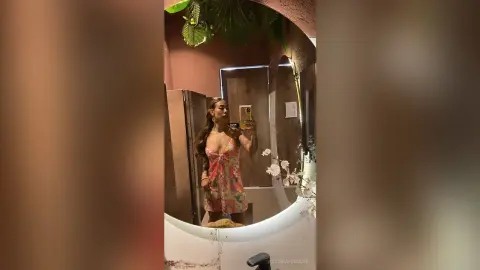 VictoriaFontaine poses in a floral sundress while taking a mirror selfie in a room with tropical plants and warm lighting.