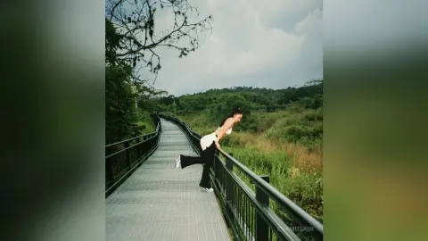 Young woman in a white crop top leaning over a metal railing on a scenic nature walkway under a cloudy sky.