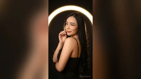 Smiling woman in a black sleeveless top posing against a glowing ring light in a dark studio setting.