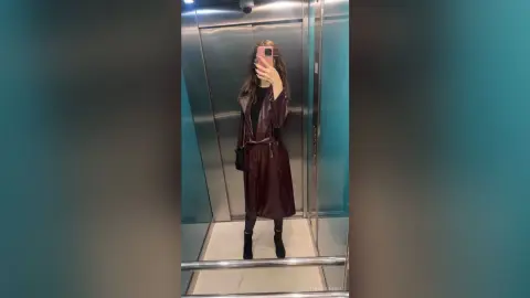 Stylish woman in a long brown leather trench coat taking a mirror selfie in a modern elevator.
