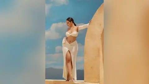 Elegant woman in a white off-the-shoulder maxi dress posing against a desert landscape under a blue sky.