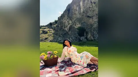 Woman in floral summer dress relaxing on a picnic blanket near rocky cliffs in a sunny meadow.