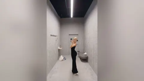Blonde woman in a sleek black evening gown taking a mirror selfie in a modern minimalist dressing room.