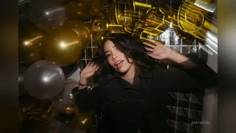 Young woman in a black coat posing joyfully amidst shimmering gold balloons in a festive party setting.