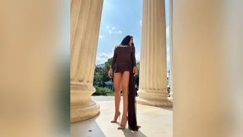 NicoleBella stands between two large stone columns wearing a tight brown long-sleeved mini dress and high heels.