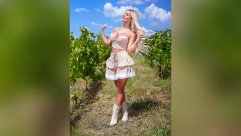 Anita stands in a vineyard wearing a tan lace bra and matching mini skirt with long blonde hair blowing in the wind.