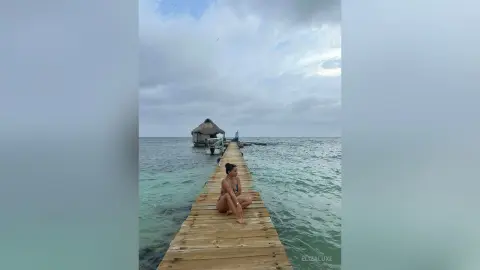 Woman in a bikini sitting on a wooden pier overlooking a calm ocean under a cloudy sky.