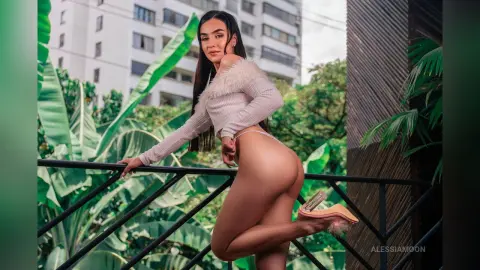 Young woman in a white long-sleeve crop top posing on a tropical balcony with lush green plants.