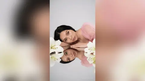 Elegant woman in a pink sheer blouse resting her head near white lilies in a soft, dreamy aesthetic.