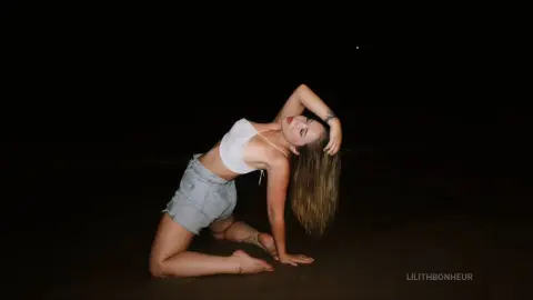 Young woman in white crop top and denim shorts posing on a dark beach at night.