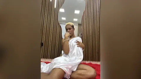 Young woman in a flowing white satin dress sitting in a modern room with wood slat walls.