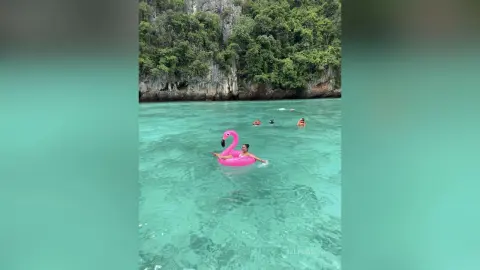 Young woman relaxing on a pink flamingo float in turquoise tropical ocean water near lush green cliffs.