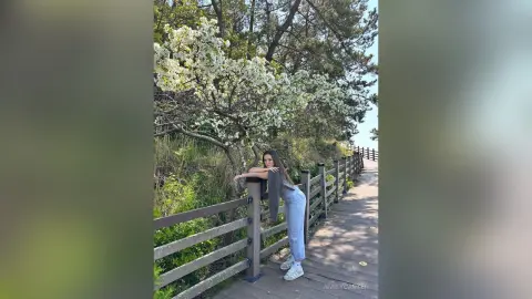 Young woman in light blue jeans and white sneakers leaning on a wooden boardwalk under blooming white flowers.