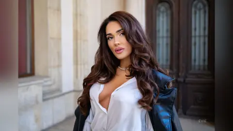 Beautiful woman in a white blouse and black leather jacket posing outdoors near a classic stone building.