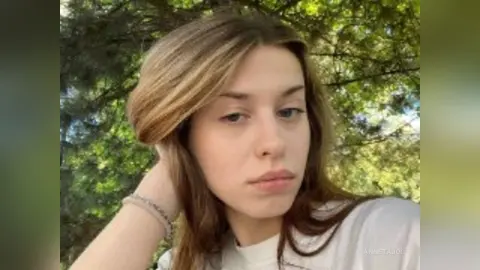 Young woman with long brown hair wearing a white t-shirt in a sunny outdoor garden setting.