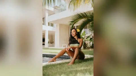 Young woman in a black bikini sitting on grass by a luxury villa pool in a tropical summer setting.