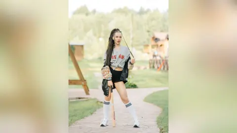 Young woman in a graphic t-shirt and black shorts holding a baseball bat in a sunny outdoor park setting.