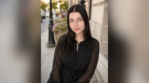 Young woman with long dark hair wearing a black polka dot blouse standing on a sunny city street.