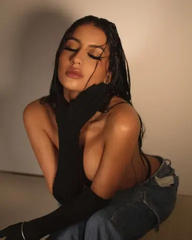 Young woman with wet hair in a black top and denim jeans posing against a neutral wall in soft lighting.
