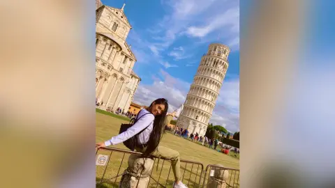 LucyMartin stands in front of the Leaning Tower of Pisa wearing a white long-sleeve shirt and tan pants.