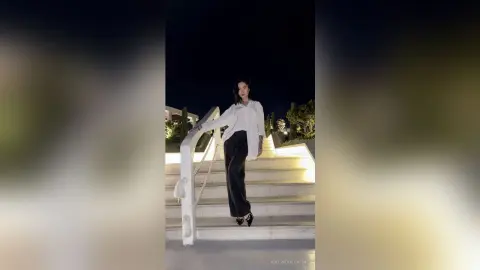 Stylish woman in a white button-down shirt and black trousers posing on white stairs at night.