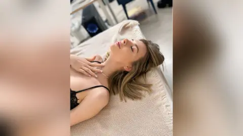 Relaxed blonde woman in black lace lingerie lying on a white textured bed in a bright, serene bedroom.