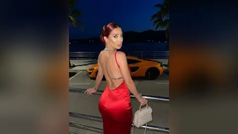DeviNessa poses with her back turned, wearing a red satin dress that exposes her back and a tattoo, standing next to an orange sports car.