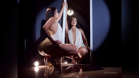MartinaBilf poses nakedly in a white top while squatting next to a pole, looking into a mirror in a dark studio setting.