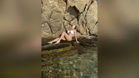 Fit woman in white bikini relaxing on coastal rocks by clear turquoise ocean water under bright sunlight.