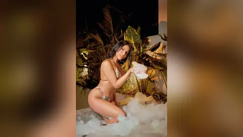 Smiling woman in a patterned bikini relaxing in a bubbly foam bath surrounded by tropical plants at night.