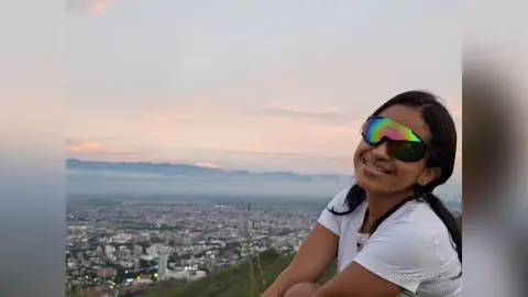 Smiling woman in white t-shirt and sunglasses enjoying a scenic sunset view over a sprawling city landscape.