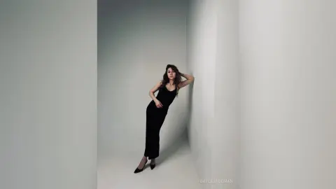 Elegant woman in a black jumpsuit leaning against a white wall in a minimalist studio setting.