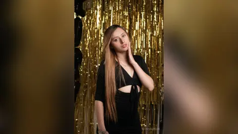 ArinnaGreen poses with long blonde hair, wearing a black cutout top against a shiny gold tinsel background.