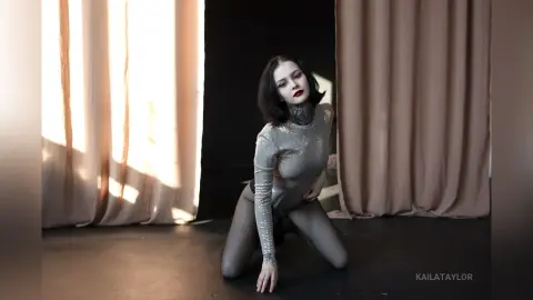 Gothic woman in a shimmering silver bodysuit posing in a moody studio setting with dramatic lighting.