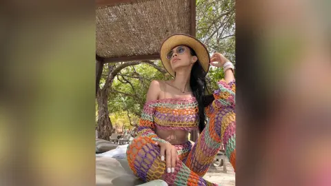 LucyMartin wears a colorful crochet crop top and matching skirt while sitting outdoors under a thatched roof.
