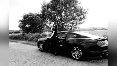 Stylish woman in black outfit sitting in a dark coupe car on a rural road under large trees.