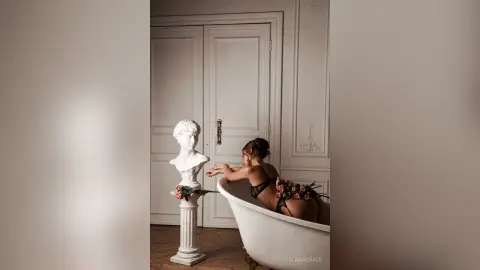 Elegant woman in black lingerie sitting in a white bathtub next to a classical bust in a bright, minimalist room.