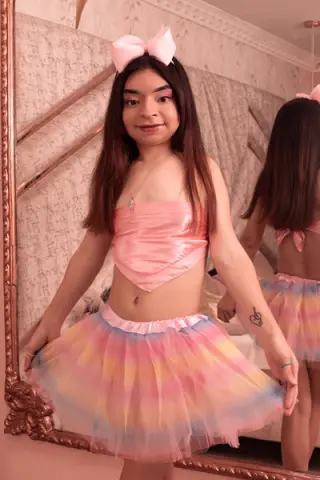 Young woman in a pink satin crop top and pastel rainbow tutu posing in front of a vintage mirror.