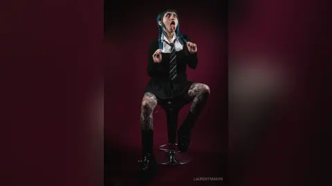 Edgy woman with blue hair in a dark schoolgirl outfit sitting on a stool with a playful expression.