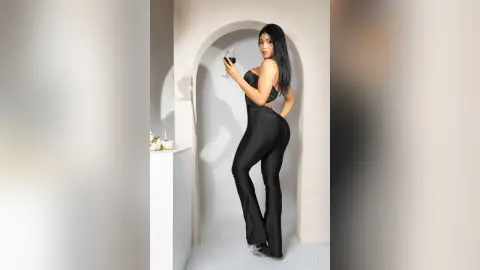 Elegant woman in a black jumpsuit holding a wine glass in a minimalist white arched interior.