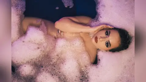 Glamorous woman with red lipstick relaxing in a dreamy, bubbly bubble bath with soft pink lighting.