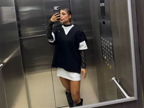 Trendy woman in a black oversized vest and white mini dress posing in a modern elevator mirror.