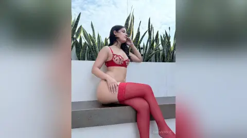 Young woman in red lace lingerie and stockings sitting on a white ledge against a tropical plant background.