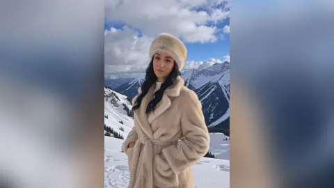 Woman in a cozy cream faux fur coat and hat smiling against a snowy mountain landscape under a blue sky.
