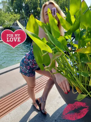 Blonde woman in a floral romper posing by lush green plants in a sunny outdoor garden setting.