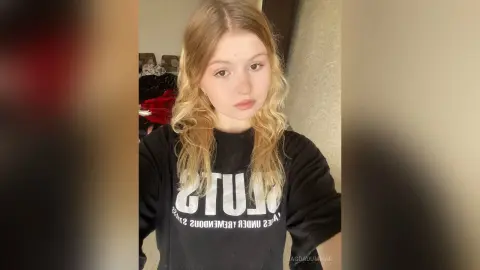 Young woman with long blonde hair wearing a black graphic sweatshirt in a cozy indoor setting.
