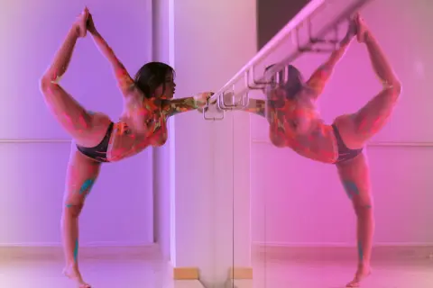 Athletic woman in colorful patterned bodysuit performing a graceful ballet stretch in a pink lit dance studio.