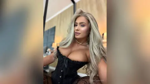 Glamorous blonde woman in a black lace corset posing in a bright, modern room.