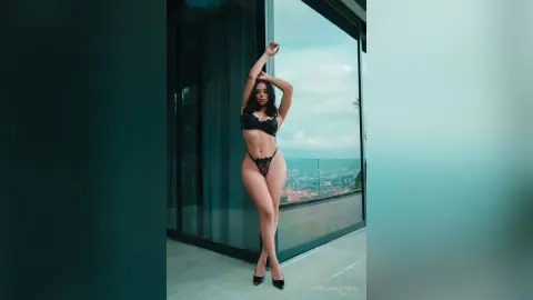 Elegant woman in black lace lingerie posing by a modern glass balcony overlooking a scenic cityscape at dusk.